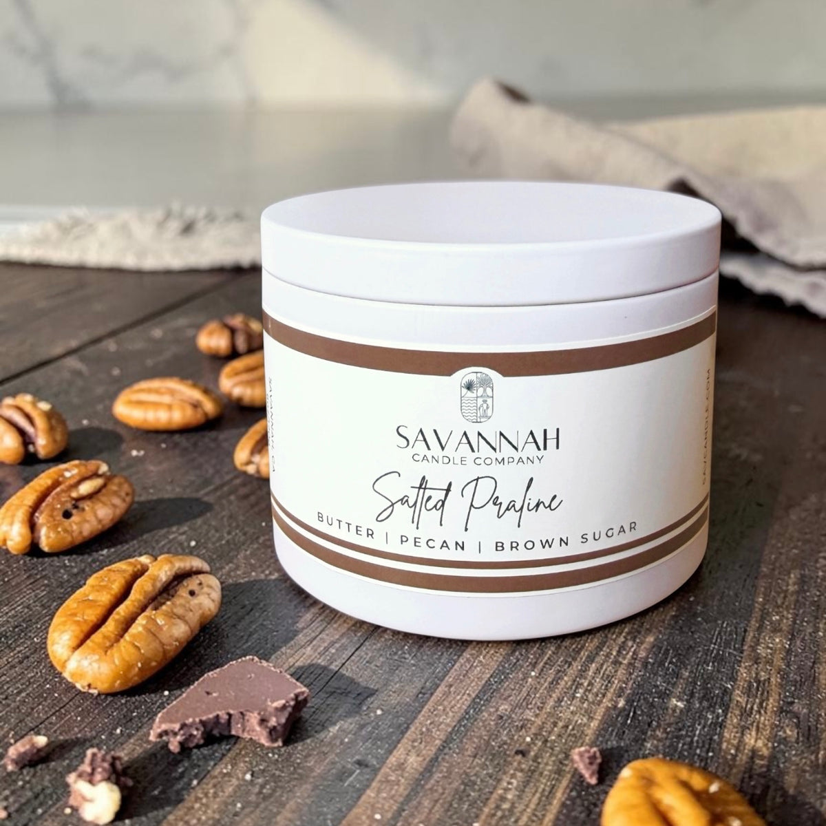 A white jar candle from Savannah Candle Company labeled 'Salted Praline' with butter, pecan, and brown sugar scent notes, styled on a dark wooden surface with scattered pecans and chocolate pieces.