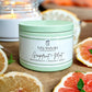 A green tin candle with a label reading 'Savannah Candle Company, Grapefruit + Mint, Peach Mint'. The candle is placed on a wooden surface with sliced grapefruits and peaches around it, and a cup with a candle inside is visible in the background.