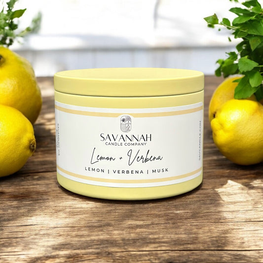A yellow tin container labeled 'Savannah Candle Company Lemon Verbena' with lemon slices and green foliage in the background.