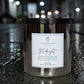 A metallic silver jar candle labeled 'Savannah Midnight' sits on a wet street at night with blurred lights in the background, showing its reflection on the pavement.