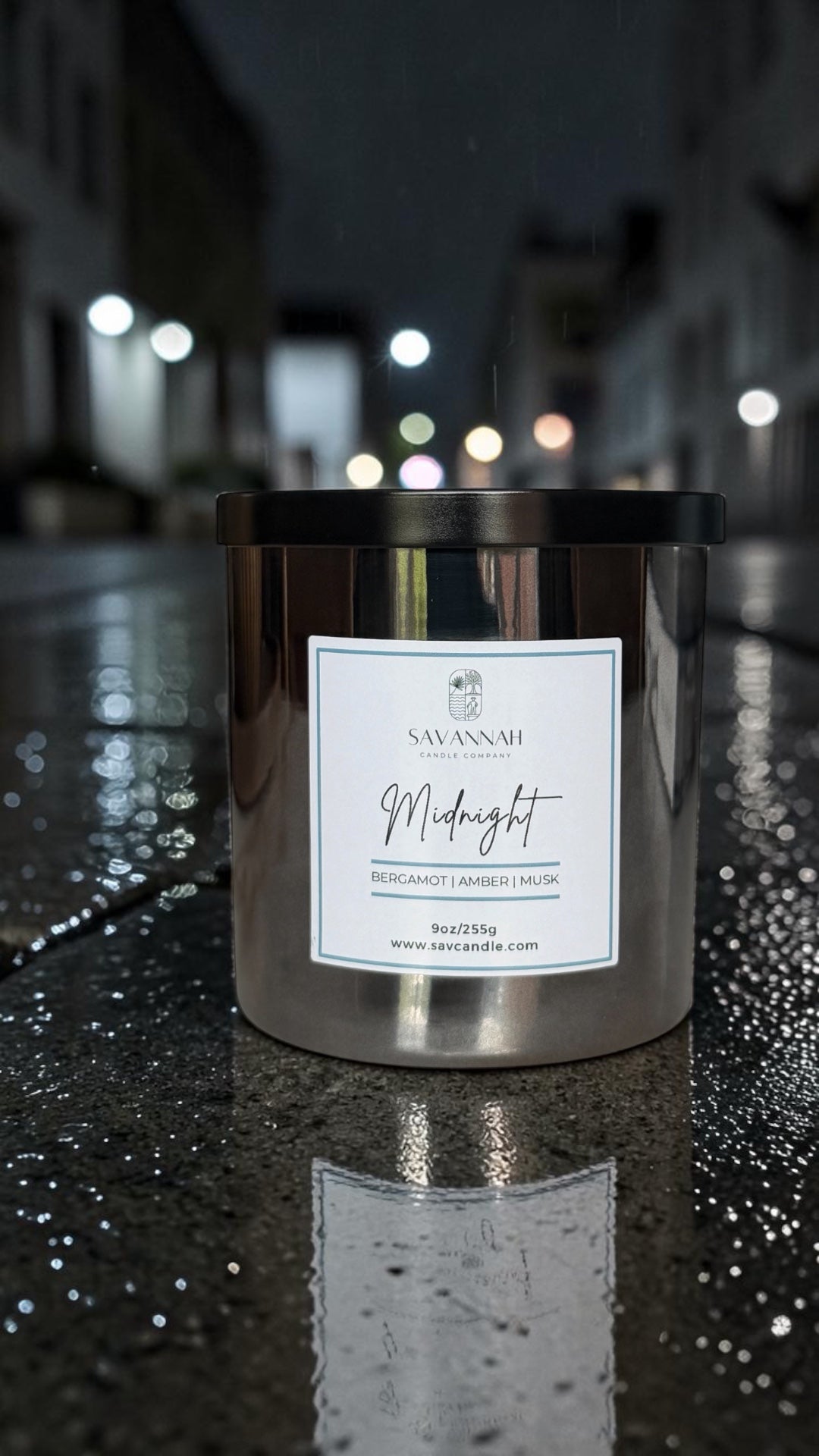 A metallic silver jar candle labeled 'Savannah Midnight' sits on a wet street at night with blurred lights in the background, showing its reflection on the pavement.