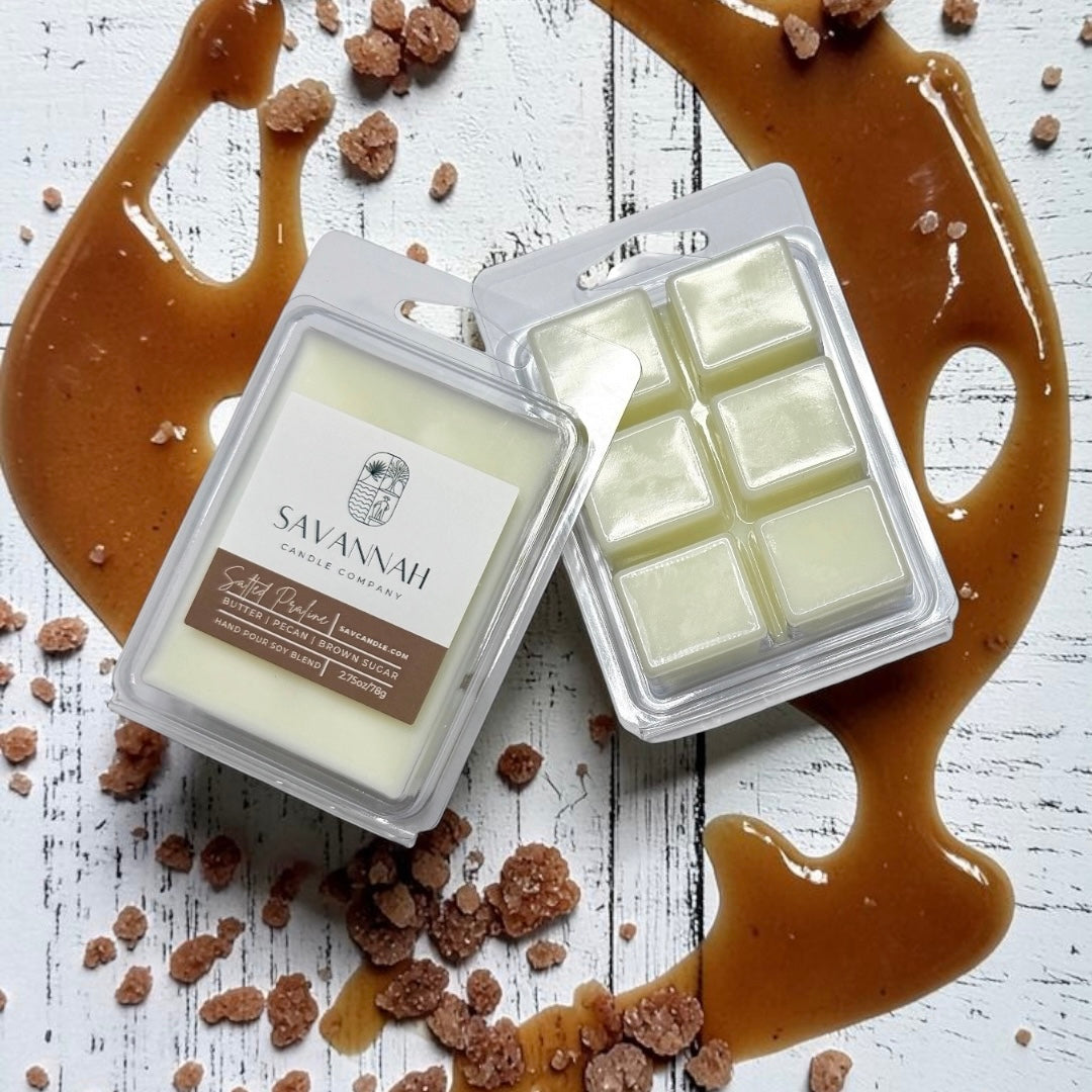Two wax melts packages labeled Savannah Candle Co. on a white distressed wood surface with caramel drizzle and brown sugar crumbs. The fragrance is salted praline.