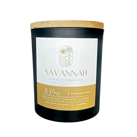 A matte black candle jar with a gold lid, labeled 'SAVANNAH CANDLE COMPANY | MOSS | EUCALYPTUS | CYPRUS | CLOVER', set against a plain background.