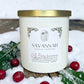 A white candle jar with a gold lid and a front label reading SAVANNAH CANDLE COMPANY, styled outdoors in snow with holly leaves and red berries around it.