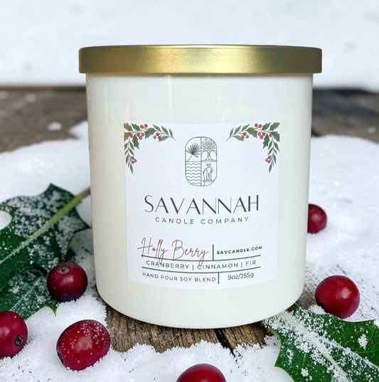 A white candle jar with a gold lid and a front label reading SAVANNAH CANDLE COMPANY, styled outdoors in snow with holly leaves and red berries around it.