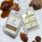 Two wax melts packages from Savannah Candle Company on a white rustic background, surrounded by brown sugar, caramel, and pecans. One package is closed with a white and brown label, and the other is open showing pale yellow wax cubes. The fragrance is salted praline