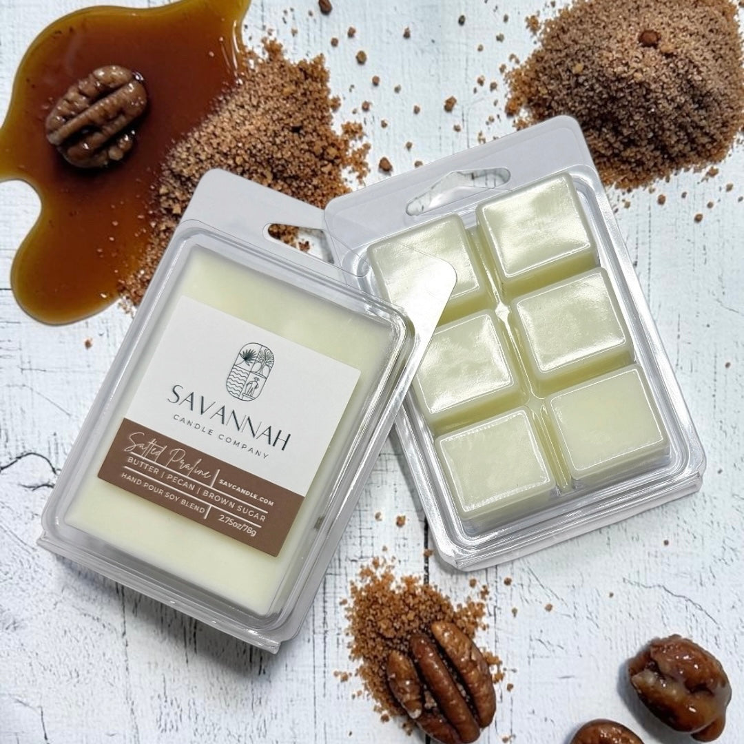 Two wax melts packages from Savannah Candle Company on a white rustic background, surrounded by brown sugar, caramel, and pecans. One package is closed with a white and brown label, and the other is open showing pale yellow wax cubes. The fragrance is salted praline