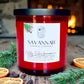 A red candle jar with a black lid and a white label reading Savannah Candle Company, styled on a rustic wooden surface with evergreen branches, dried orange slices, and a blurred fireplace in the background. The name of the fragrance is Holiday Embers