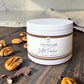 A white jar candle from Savannah Candle Company labeled 'Salted Praline' with butter, pecan, and brown sugar scent notes, styled on a dark wooden surface with scattered pecans and chocolate pieces.