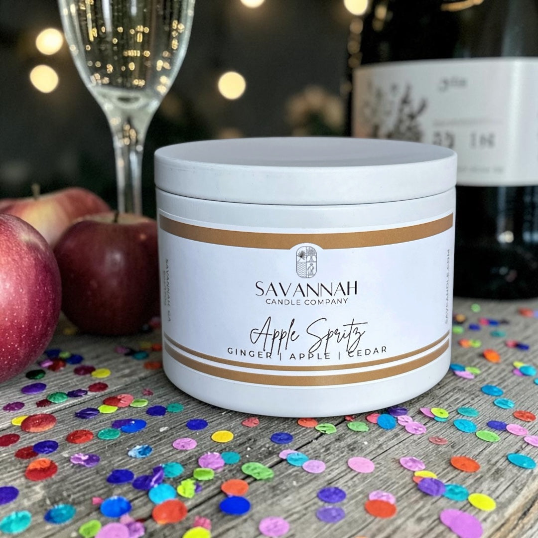 A white jar candle labeled 'Savannah Candle Company Apple Spritz' sits on a rustic wooden surface scattered with colorful confetti, with apples, a champagne flute, and blurred bottles in the background.