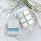 Two packages of Savannah Single Purpose Wax melts on white sand with beach grass. One package is closed, showing a white wax melt with a light blue label, and the other is open, showing six white wax cubes in a clear clamshell.