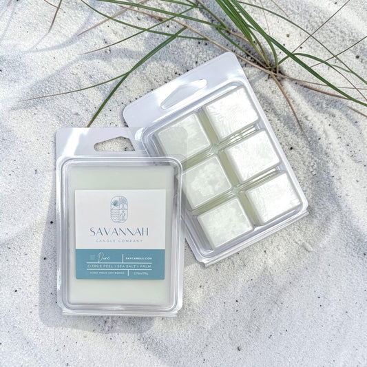 Two packages of Savannah Single Purpose Wax melts on white sand with beach grass. One package is closed, showing a white wax melt with a light blue label, and the other is open, showing six white wax cubes in a clear clamshell.