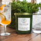 Savannah Candle Company candle on a table with a glass of iced gin and a cypress branch.  The candle fragrance is Gin and Cypress.