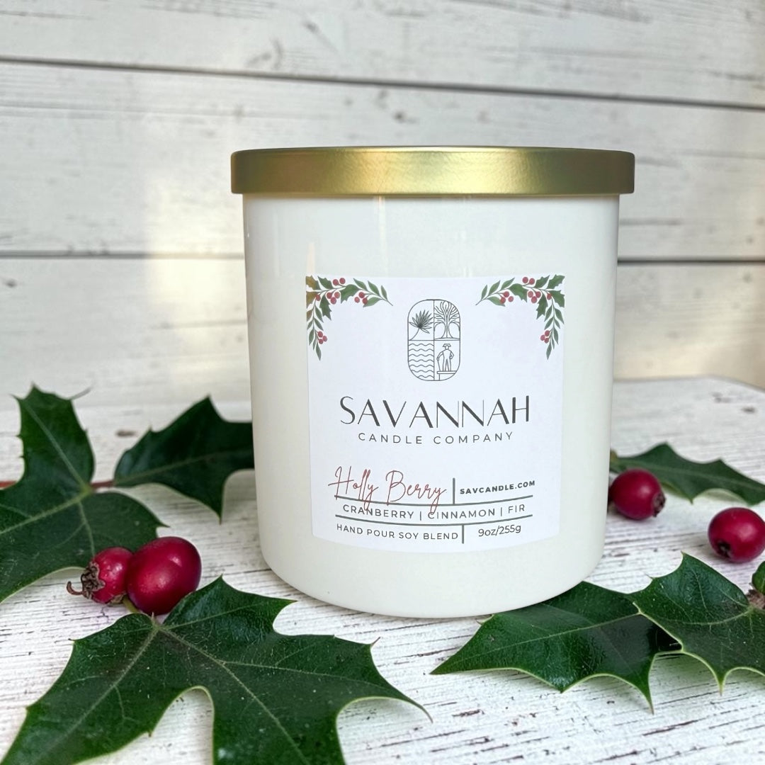 A white jar candle with a gold lid and a white label reading Savannah Candle Company, styled with green holly leaves and red berries on a white wood surface.  Fragrance is cranberry, pine, and cinnamon. 