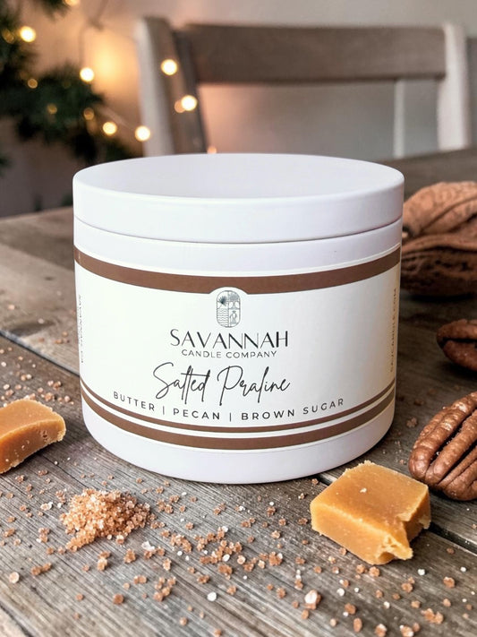 A white plastic jar of Savannah Candle Company 'Salted Praline' candle, styled on a rustic wooden table with caramel pieces, brown sugar, and pecans, with a blurred background showing warm string lights.