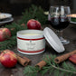 A Savannah Candle Company 'Wissal' orange and apple cider + port scented candle in a white tin with its lid leaning beside it, styled on a rustic wooden surface with red apples, cinnamon sticks, pine branches, and a glass of red wine.