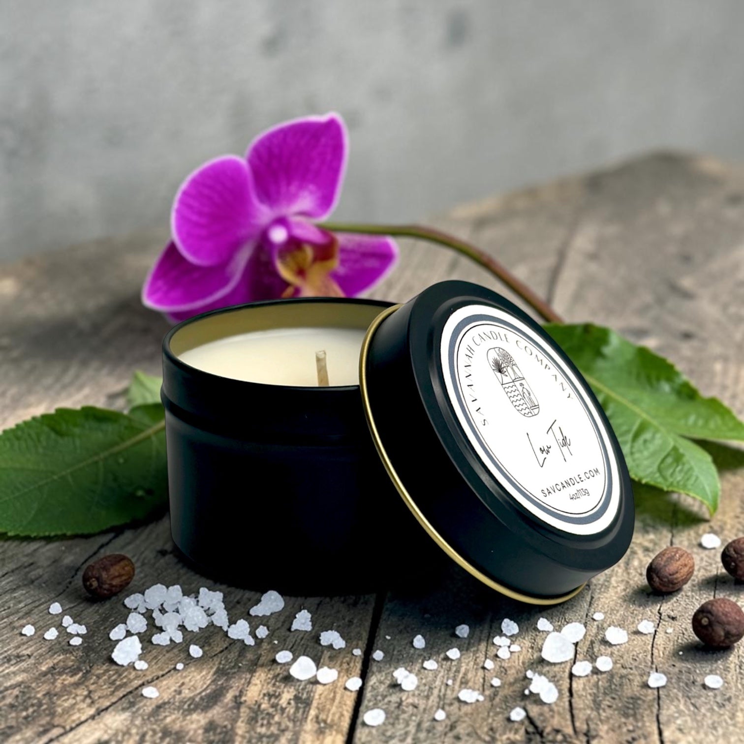 A black metal candle tin with its lid leaning beside it, filled with white wax and set on a rustic wooden surface with scattered salt crystals, coffee beans, green leaves, and a purple orchid flower. The lid label shows the brand name Lavender Creek Candle Co.