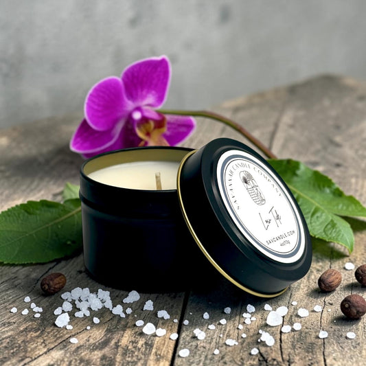 A black metal candle tin with its lid leaning beside it, filled with white wax and set on a rustic wooden surface with scattered salt crystals, coffee beans, green leaves, and a purple orchid flower. The lid label shows the brand name Lavender Creek Candle Co.