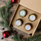 An open kraft gift box holding four small candles with gold tins and white labels, arranged on a rustic wooden surface with pine branches, red ornaments, and berries around it.  The four fragrances are Gin and Cypress, apple spritz, wassail, and salted praline.   