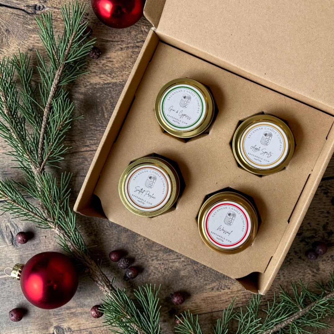 An open kraft gift box holding four small candles with gold tins and white labels, arranged on a rustic wooden surface with pine branches, red ornaments, and berries around it.  The four fragrances are Gin and Cypress, apple spritz, wassail, and salted praline.   