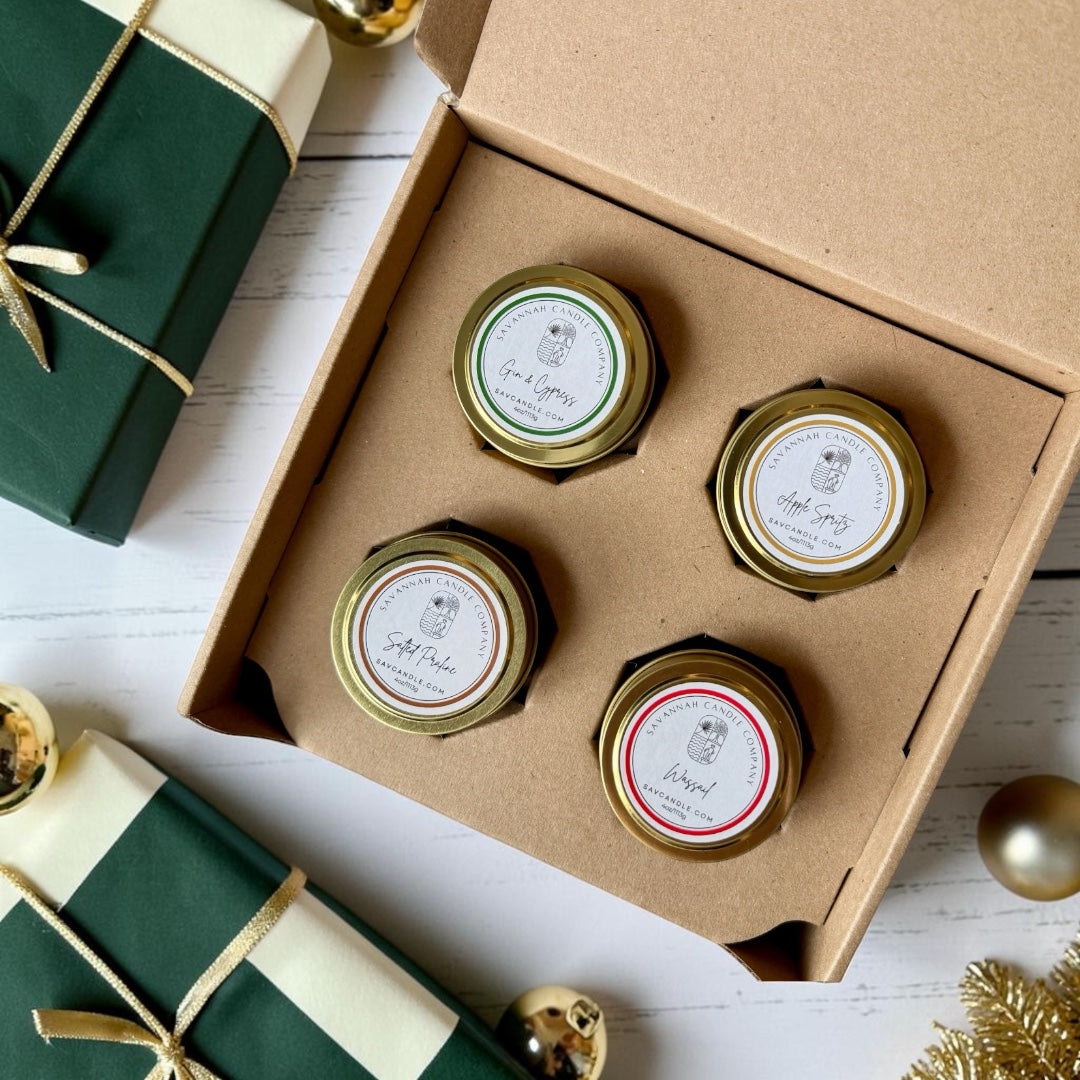 Four small candle tins with gold lids arranged in a brown kraft gift box on a white wood surface. The jars have white labels with black text and decorative borders in green, red, and gold.  The four fragrances are gin and Cypress, apple spritz, salted praline, and wassail.  
