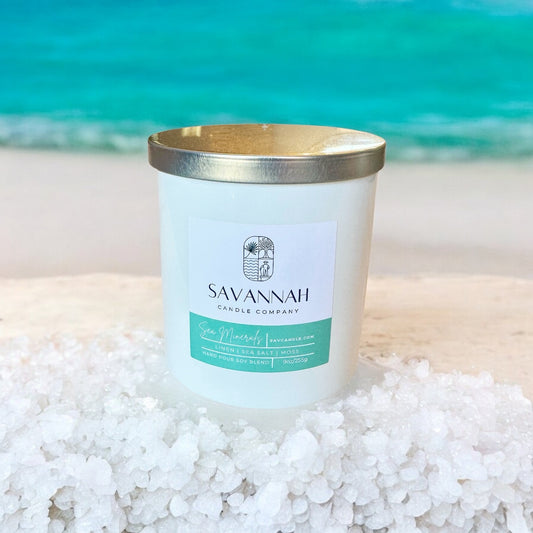 Savannah Candle Company candle on a sandy beach with ocean in the background.  The fragrance of this candle has sea salt, ocean, air, and salt water.  