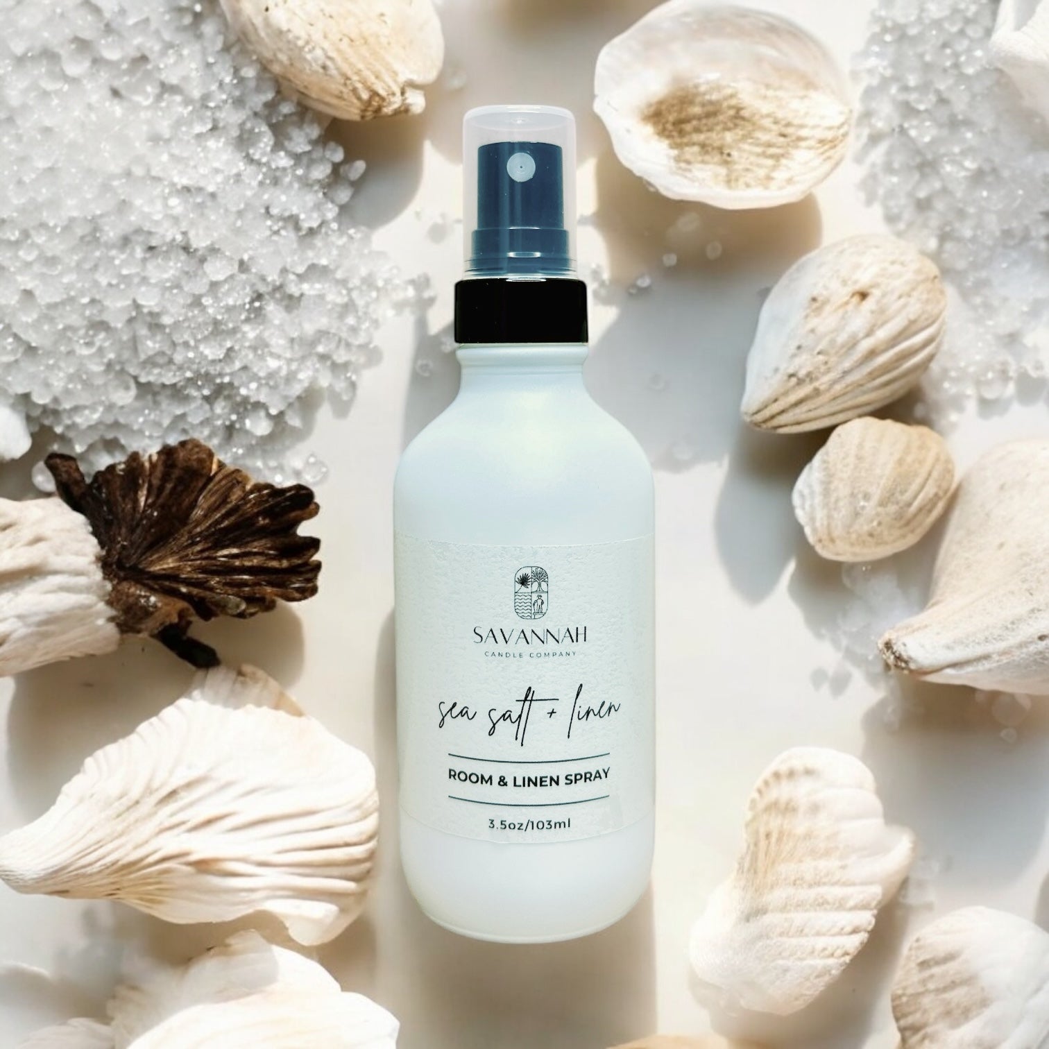 A matte white room spray bottle labeled 'SAVANNAH sea salt + linen' surrounded by seashells and white salt on a light background.