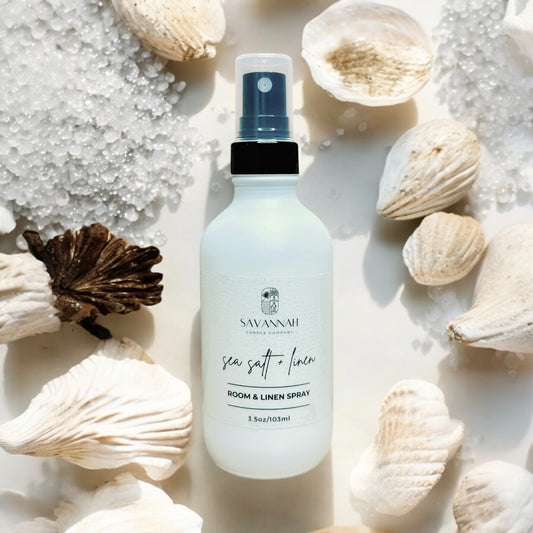 A matte white room spray bottle labeled 'SAVANNAH sea salt + linen' surrounded by seashells and white salt on a light background.