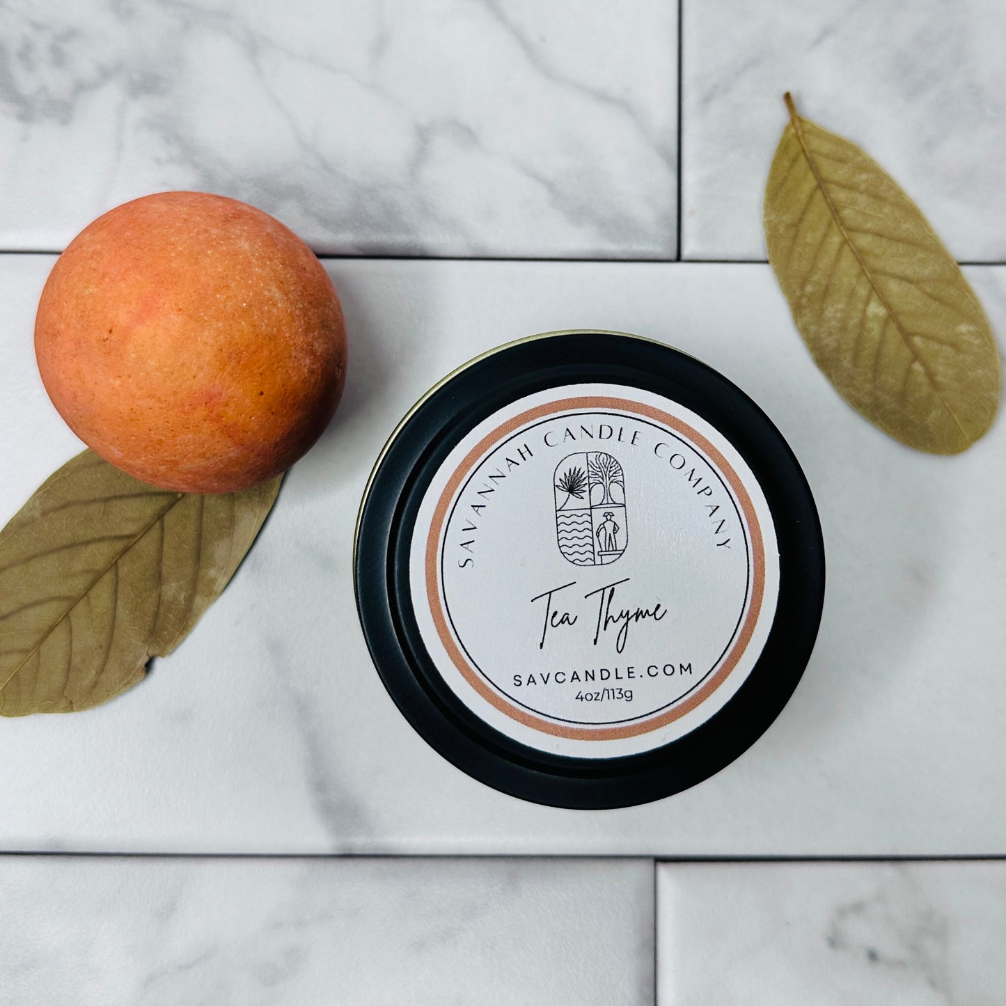 A black tin candle container with a label reading 'TEA THYME', accompanied by a lemon and a leaf on a marble surface.